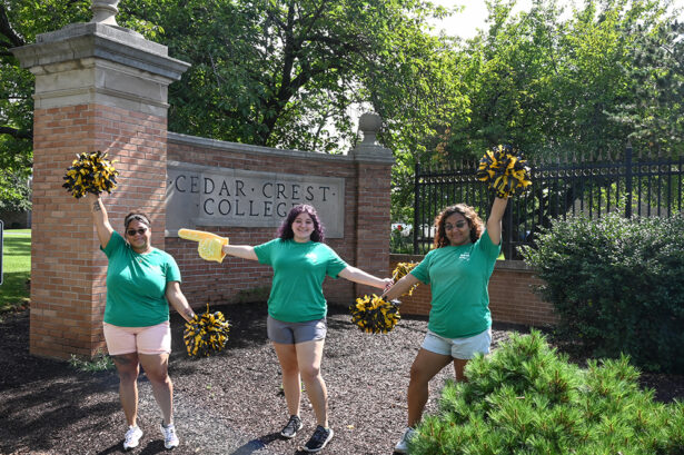 Three students in green t-shirts standing in front of South Gate with pom-poms and cheering.